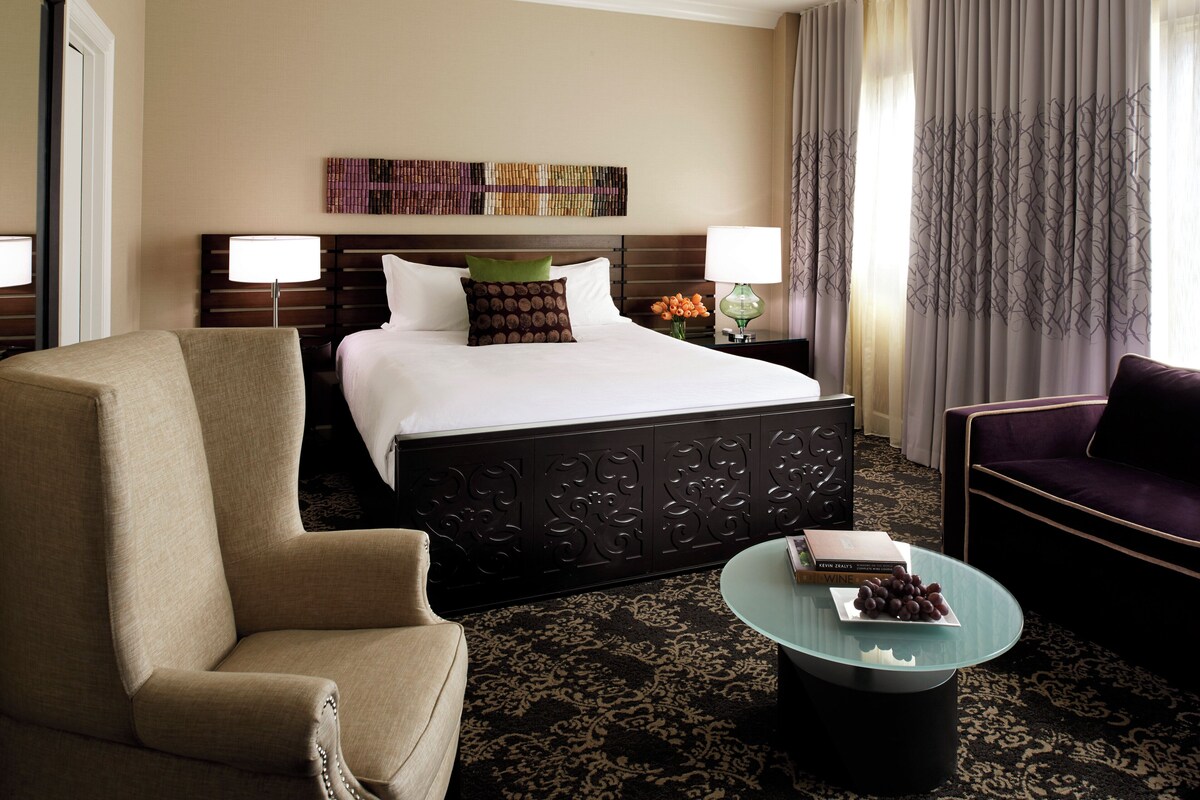 A hotel room with a large bed, two bedside lamps, a green vase with flowers, a glass coffee table with a book and grapes, and a dark patterned carpet.