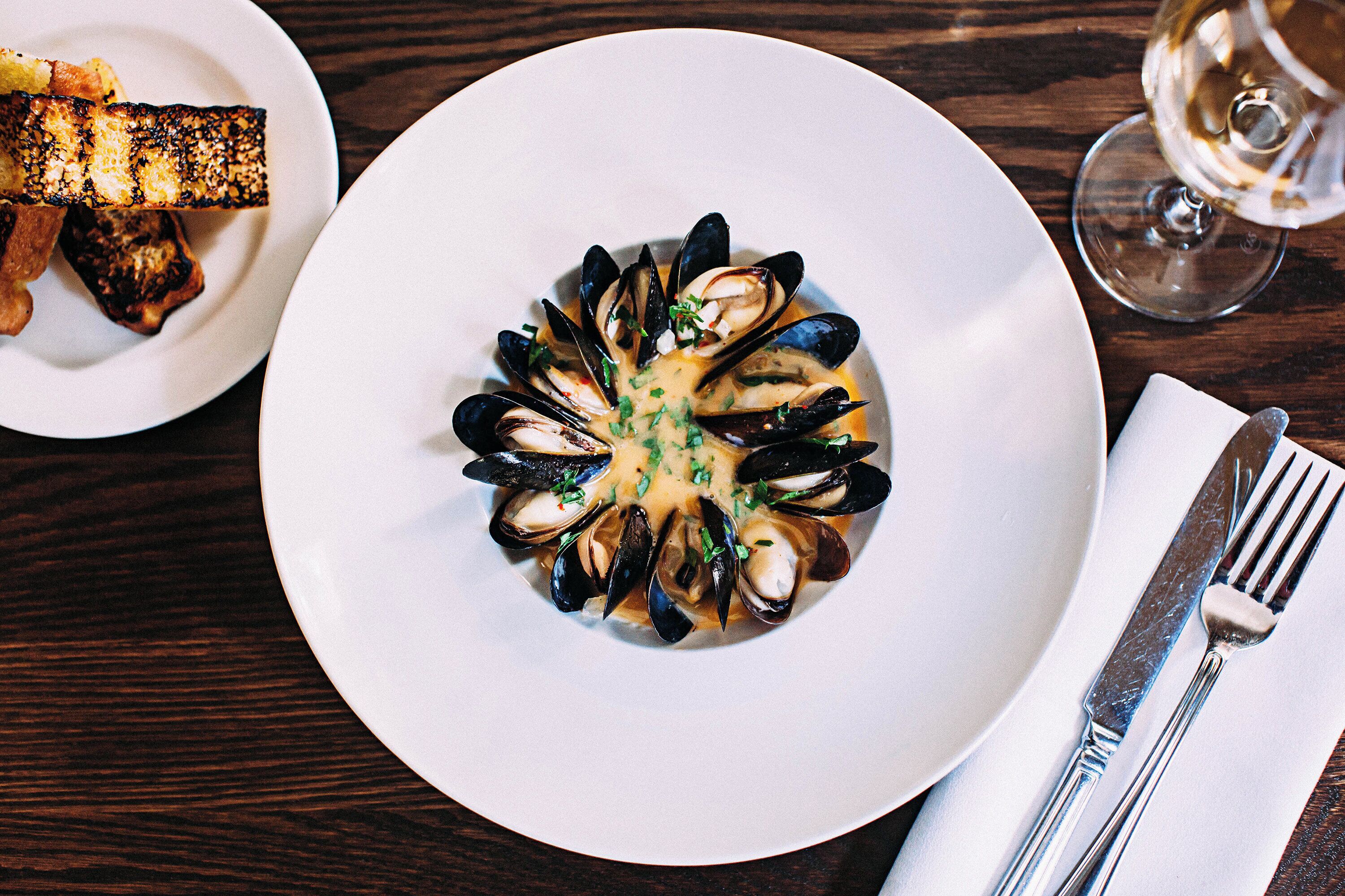 A plate of mussels with a creamy sauce, a fork, and a knife on a wooden table.