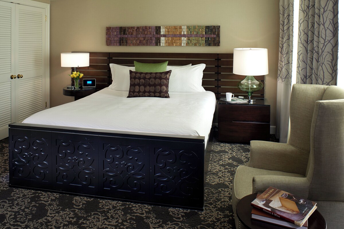 A hotel room with a large bed, two bedside tables, a chair, and a wall art piece.