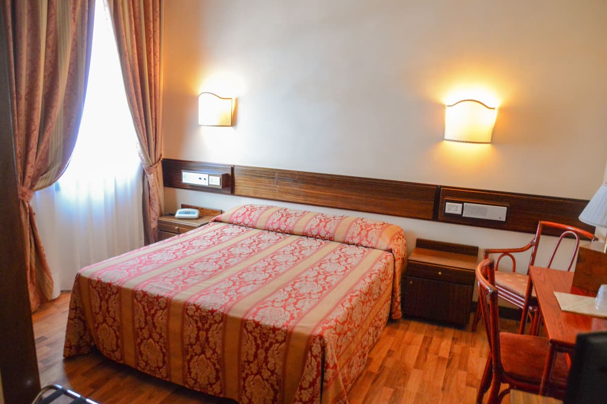 A hotel room with a bed, bedside tables, a nightstand, a wooden chair, and a table with a lamp.