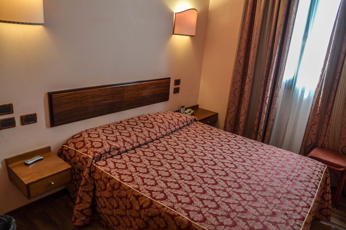 A hotel room with a large bed, bedside tables, a nightstand, a lamp, and a view of the outside through curtains.