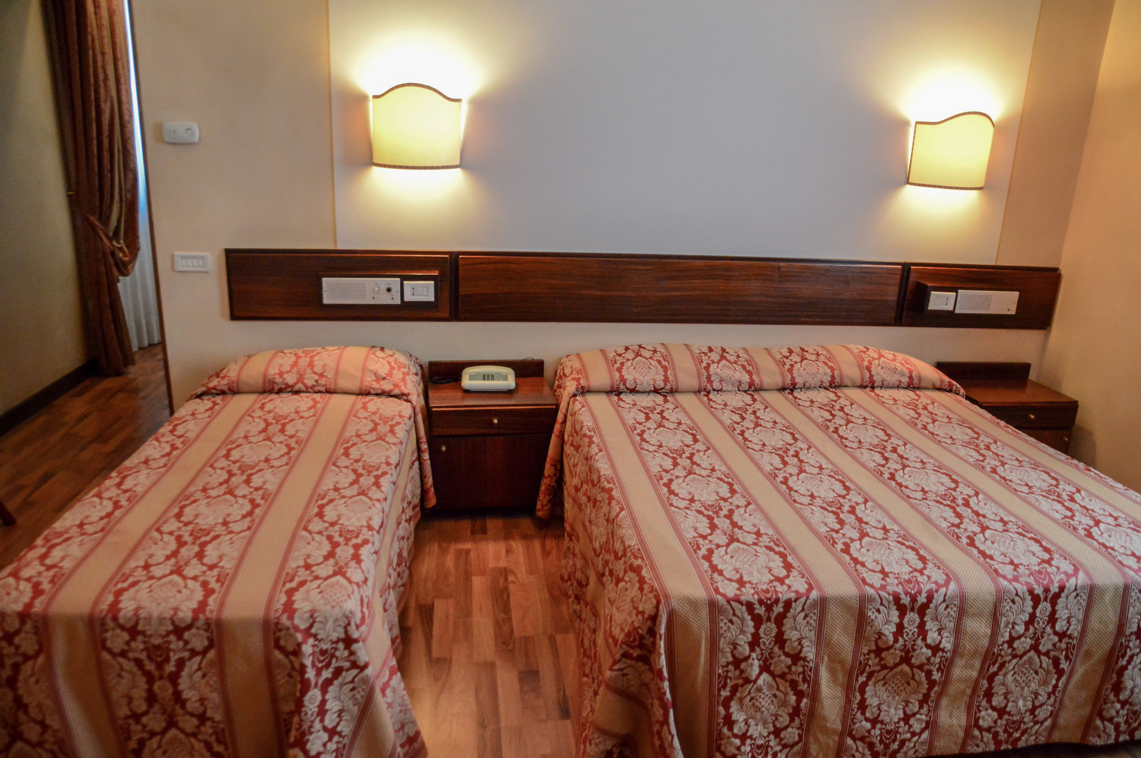 A hotel room with two beds, wooden headboards, bedside tables, and wall-mounted lamps.