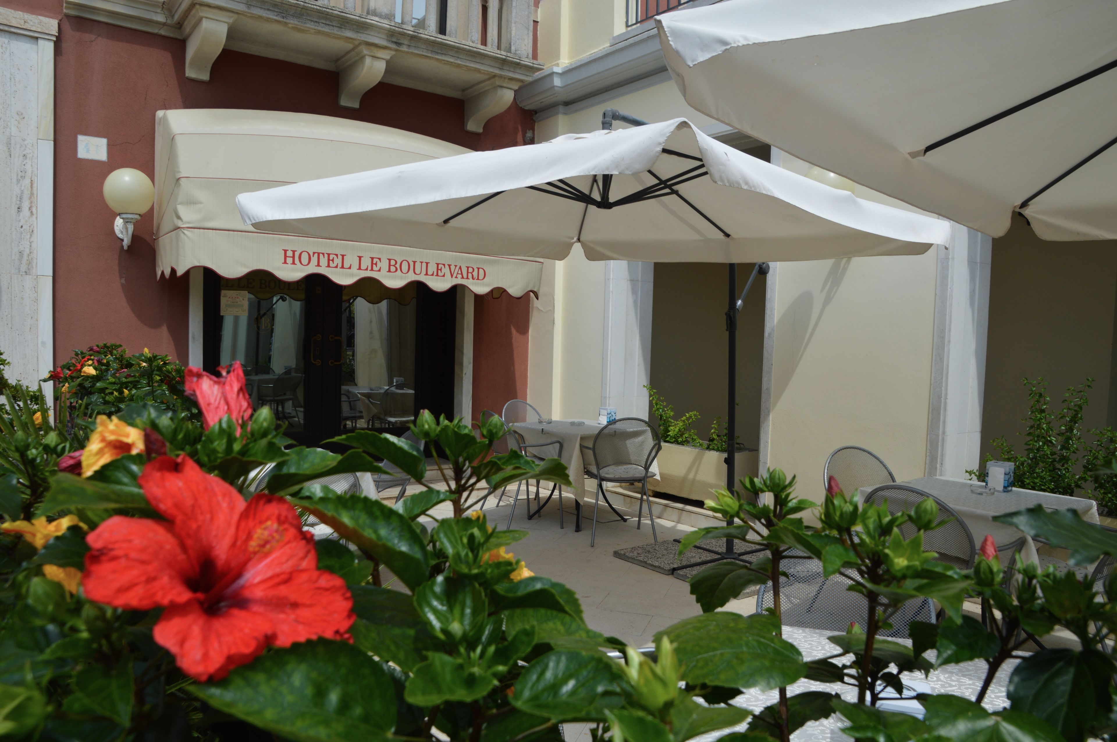 Un espace extérieur aménagé avec des tables et des chaises, un parasol blanc et un bâtiment sur lequel on peut lire l’inscription « HÔTEL LE BOULEVARD ».