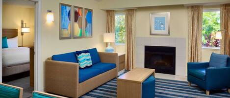 A hotel room with a bed, a blue sofa, a blue armchair, a wooden coffee table, and a fireplace.