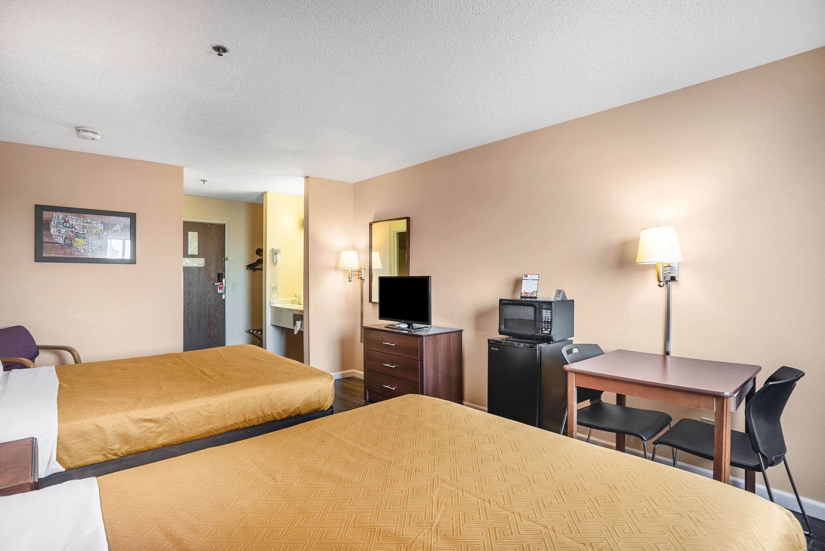 A hotel room with two beds, a desk with a chair, a television, a refrigerator, and a lamp.