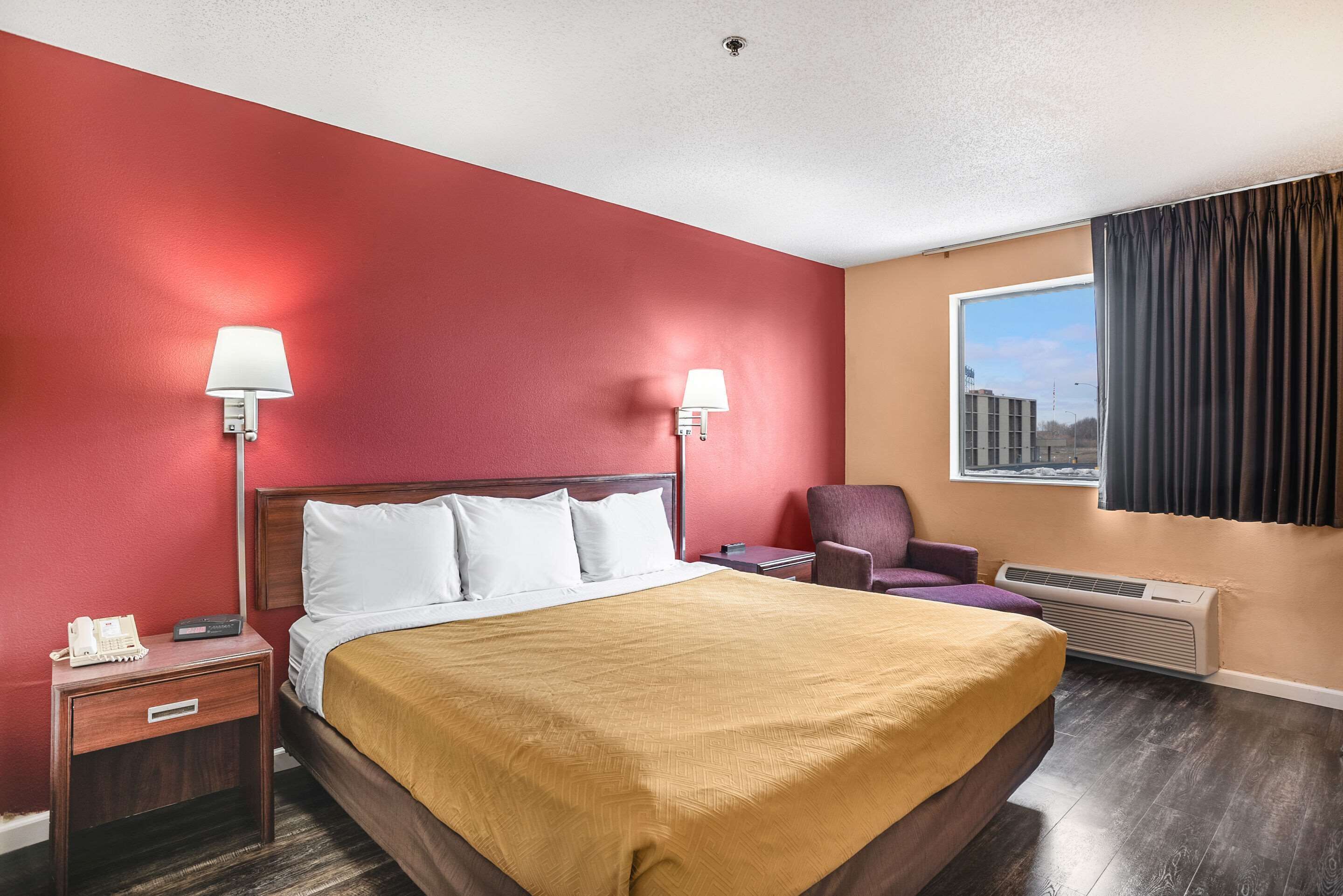 A hotel room with a large bed, two bedside lamps, a chair, a window with curtains, and a thermostat.