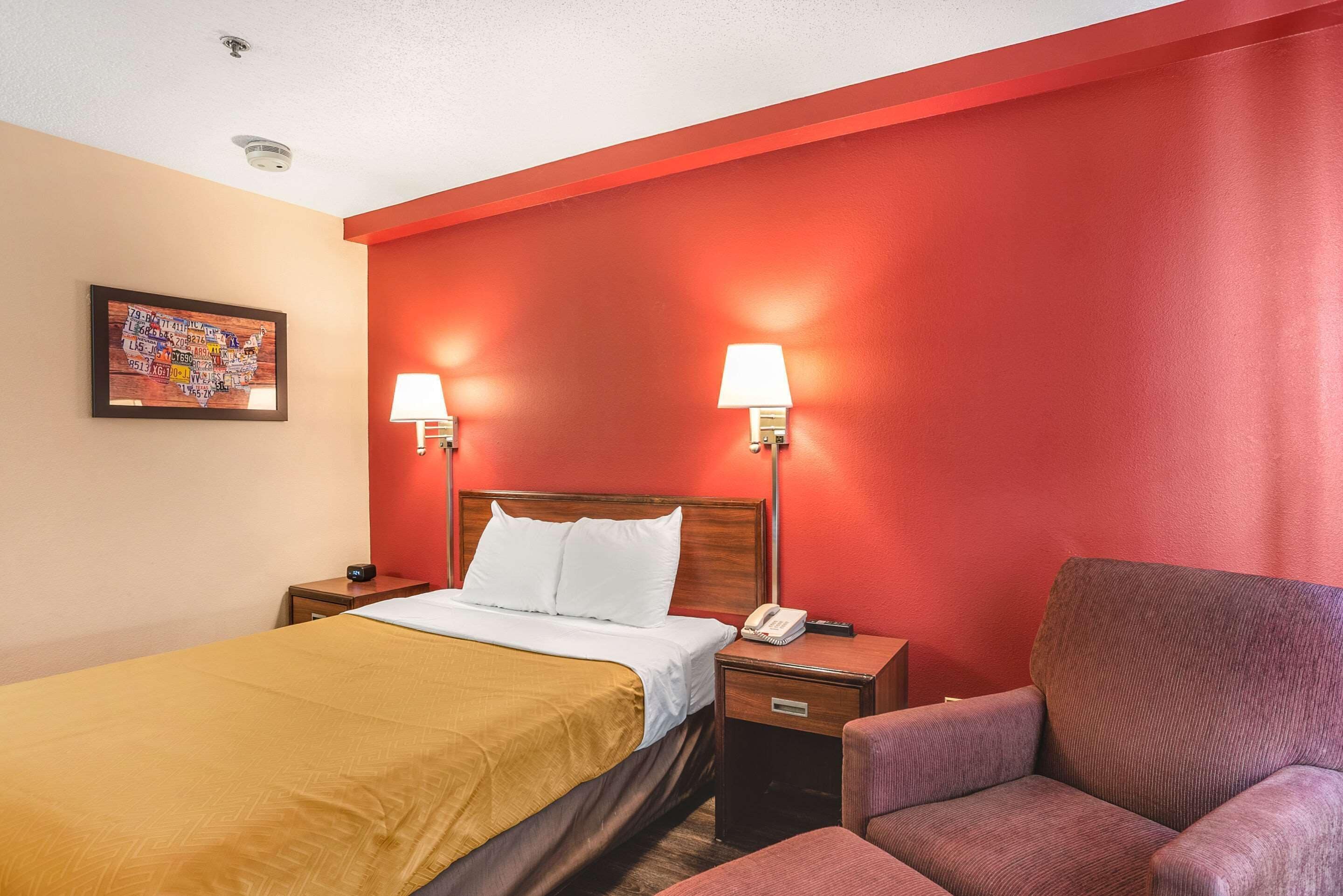 A hotel room with a bed, two bedside lamps, a chair, a nightstand with a phone, and a wall with a red accent and a framed picture.