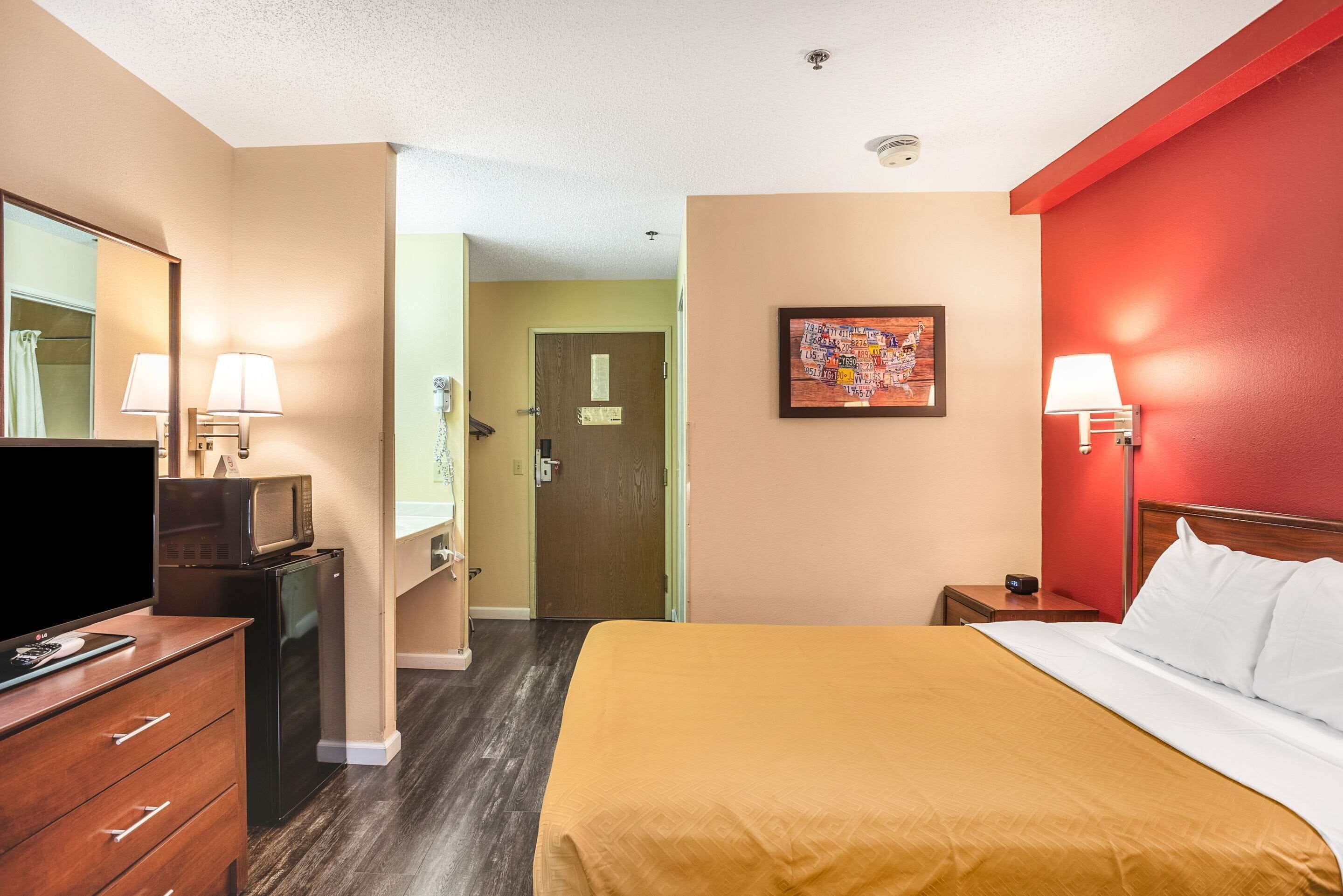 Una habitación de hotel con una cama, un televisor sobre una cómoda, un baño con lavamanos y espejo, y una puerta que da a otra habitación.