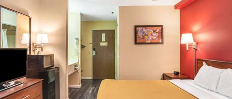 Una habitación de hotel con una cama, un televisor sobre una cómoda, un baño con lavamanos y espejo, y una puerta que da a otra habitación.