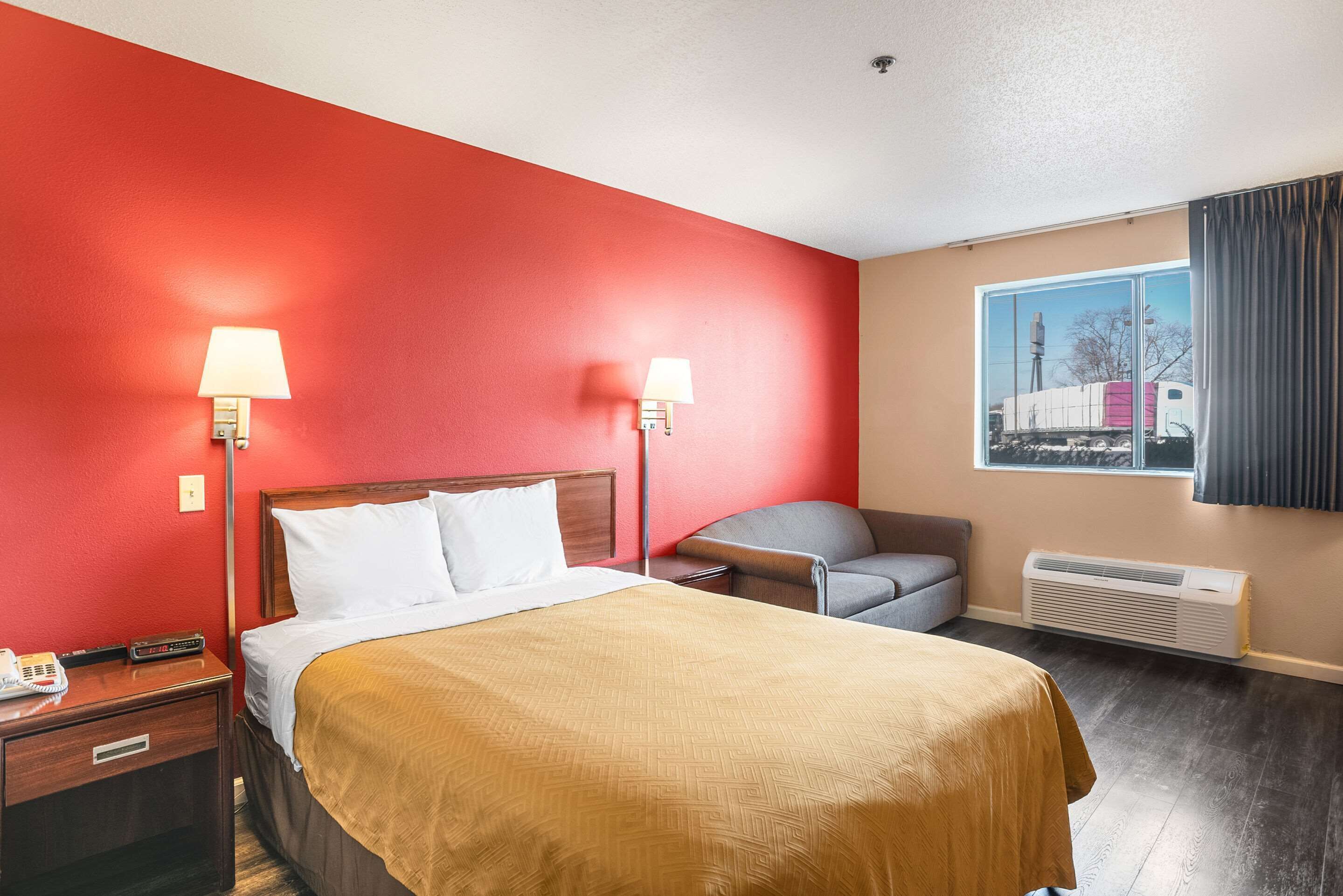 A hotel room with a red wall, a bed with white bedding, two bedside lamps, a gray armchair, and a window with a view of a building.
