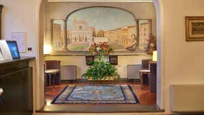 A lobby with a mural of a cityscape, a reception desk, patterned chairs, a blue carpet, and a potted plant.
