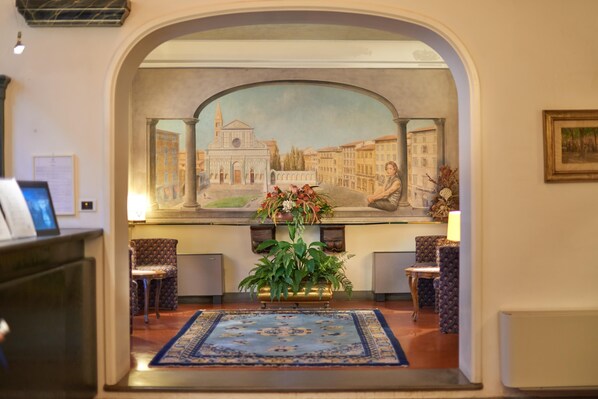 A lobby with a mural of a cityscape, a reception desk, patterned chairs, a blue carpet, and a potted plant. - Rivoli Boutique Hotel (Florence)