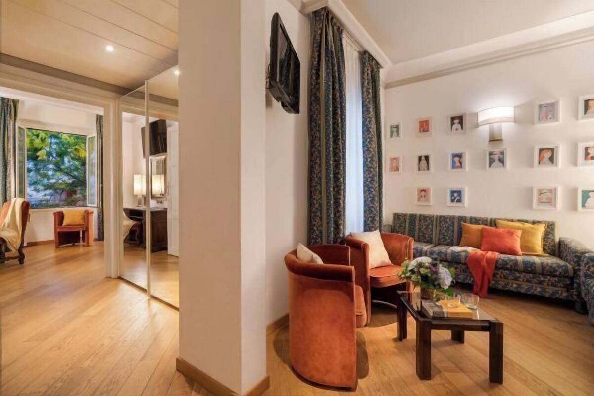 Un salon comprenant un canapé, des fauteuils, une table basse et un téléviseur. Des tableaux encadrés ornent le mur, et une fenêtre est agrémentée de rideaux.