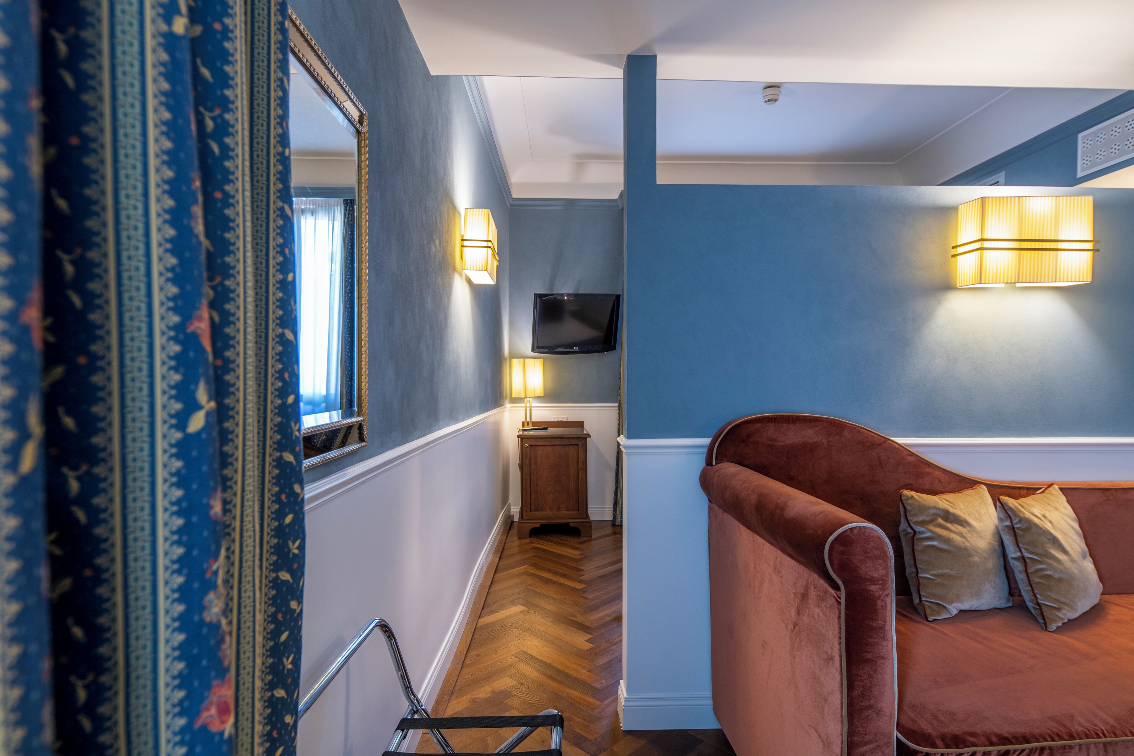 A room with a wooden floor, a brown sofa, a television mounted on the wall, and a mirror reflecting part of the room.