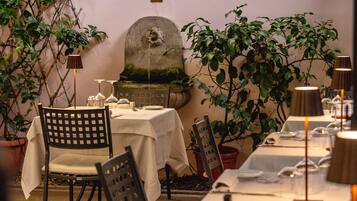 Un espace repas avec des tables préparées pour deux personnes, des chaises, des plantes en pot et une fontaine en pierre.
