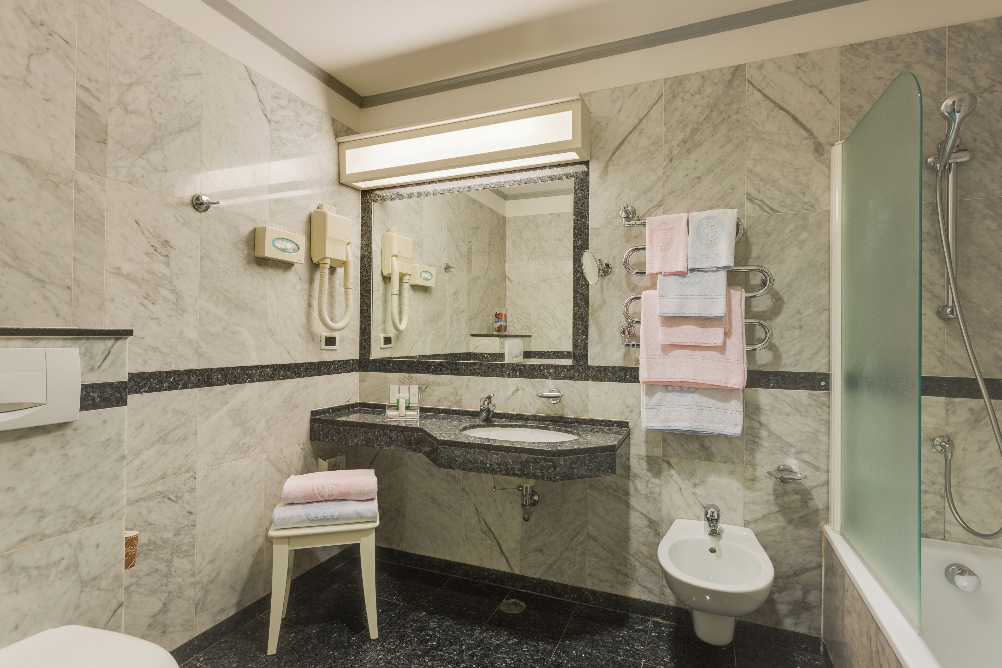 A bathroom with a marble wall, a sink, a bidet, a mirror, and a glass-enclosed shower.