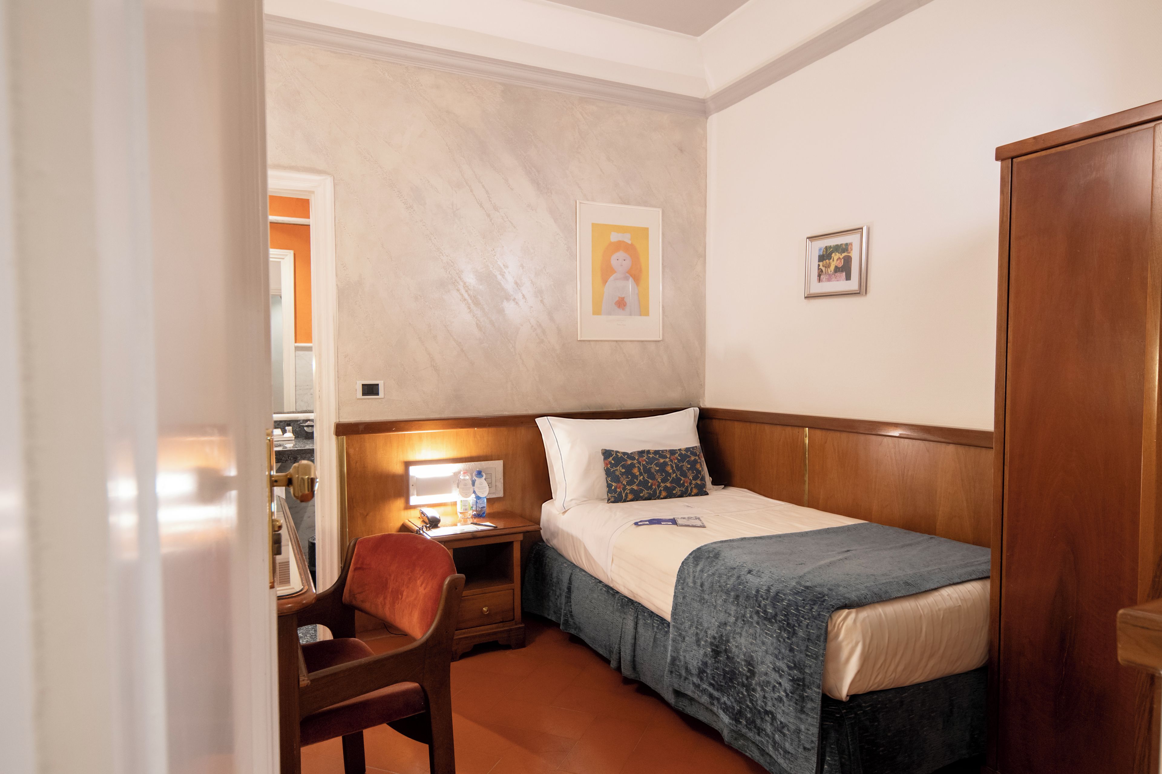 A hotel room with a bed, a desk, a chair, and two framed pictures on the wall.
