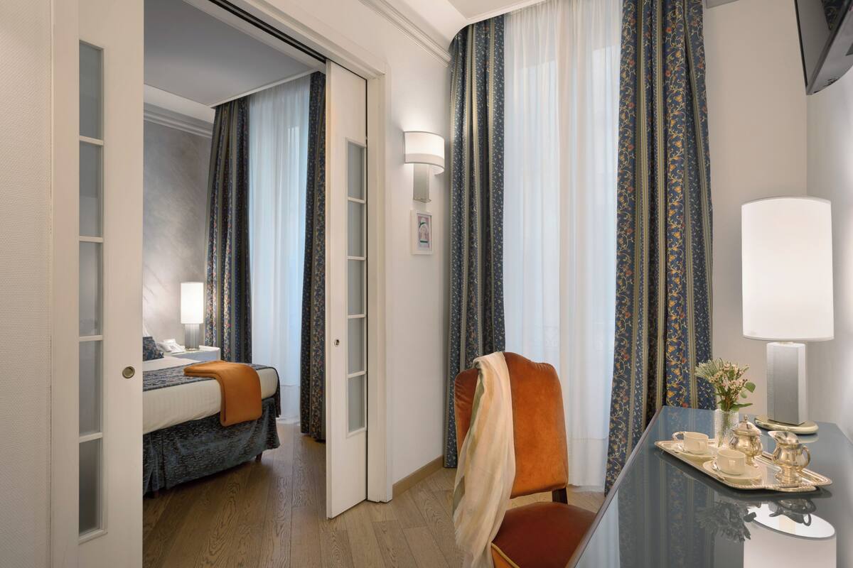 Une chambre d’hôtel avec un lit, une chaise, un bureau sur lequel se trouve un plateau avec du thé et des rideaux.