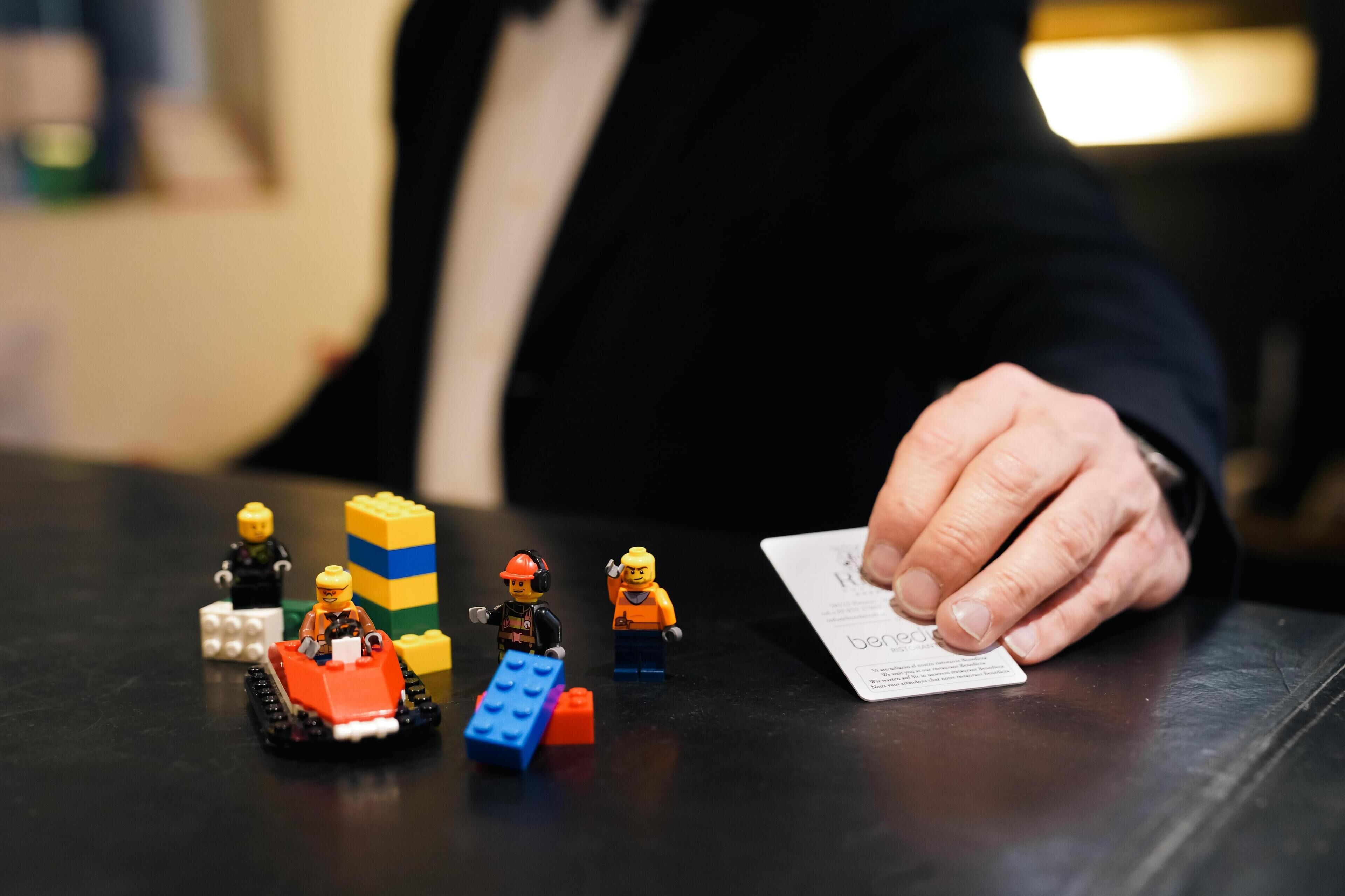 Una persona con traje negro sentada a una mesa con figuras LEGO, una tarjeta y algunos bloques de construcción.