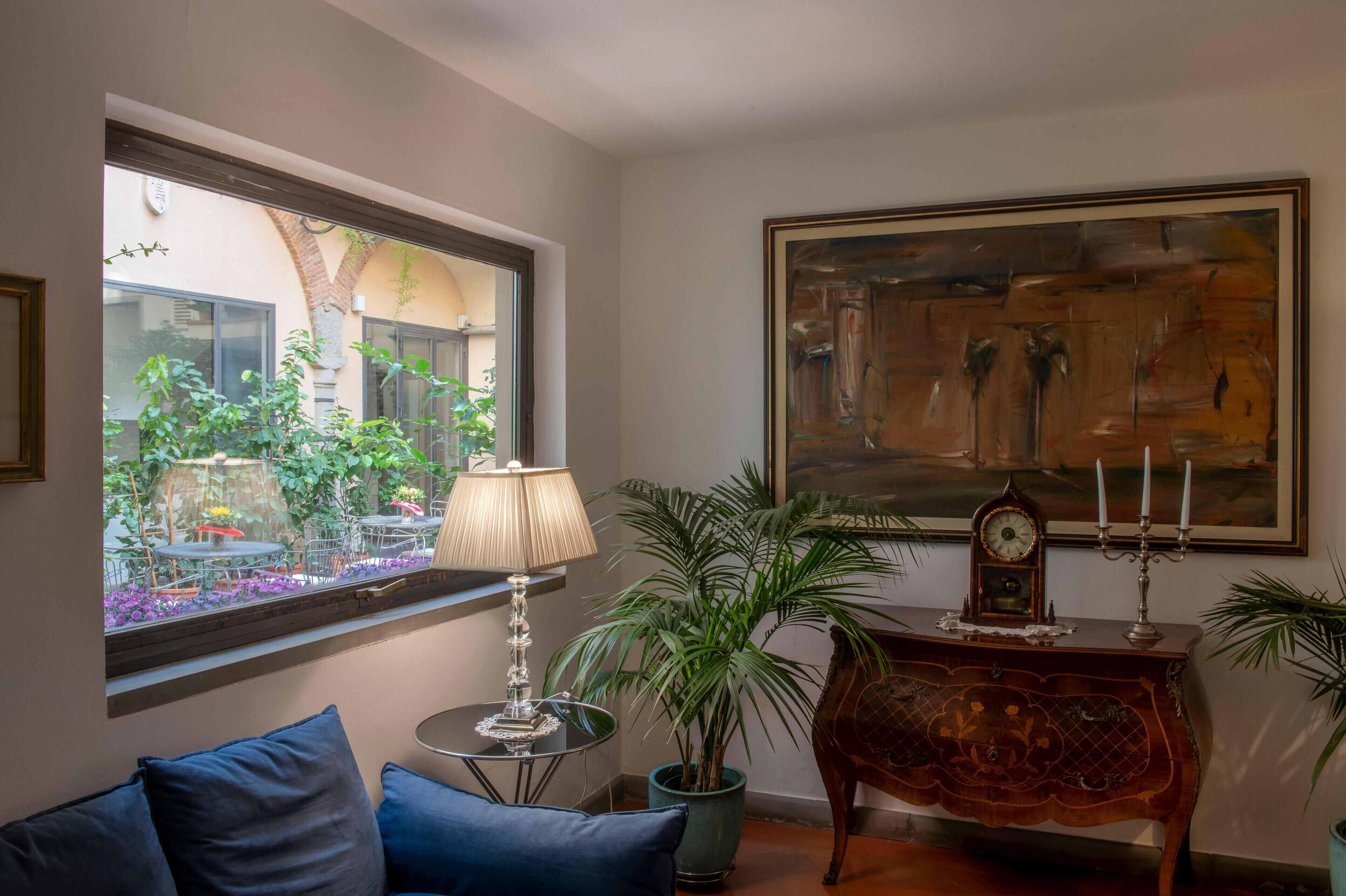 Une pièce chaleureuse avec une grande fenêtre, une plante en pot, une table avec une lampe, une horloge et un tableau encadré.