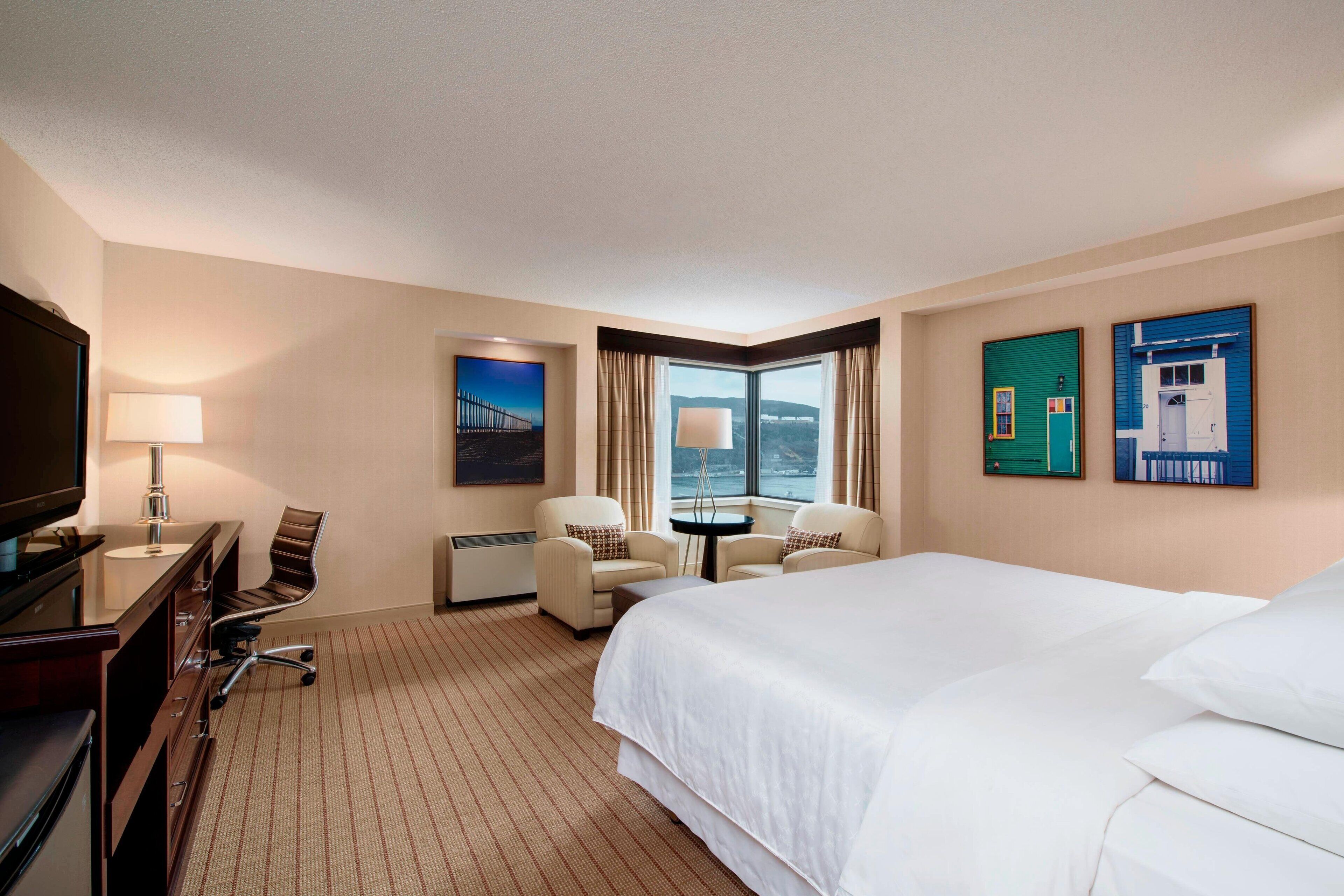 A hotel room with a large bed, a desk with a TV, two armchairs, a small table, and a window with a view.