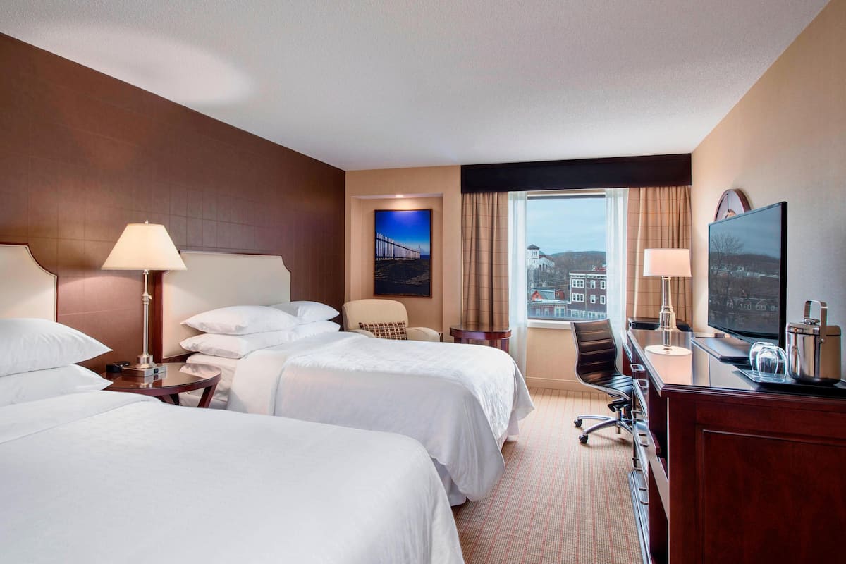 Ein Hotelzimmer mit zwei Betten, einem Schreibtisch, einem Fernseher, einem Sessel und einem Fenster mit Blick auf die Stadt.