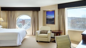Habitación de hotel con cama, ventana redonda, una silla, una mesita y vista a la ciudad.