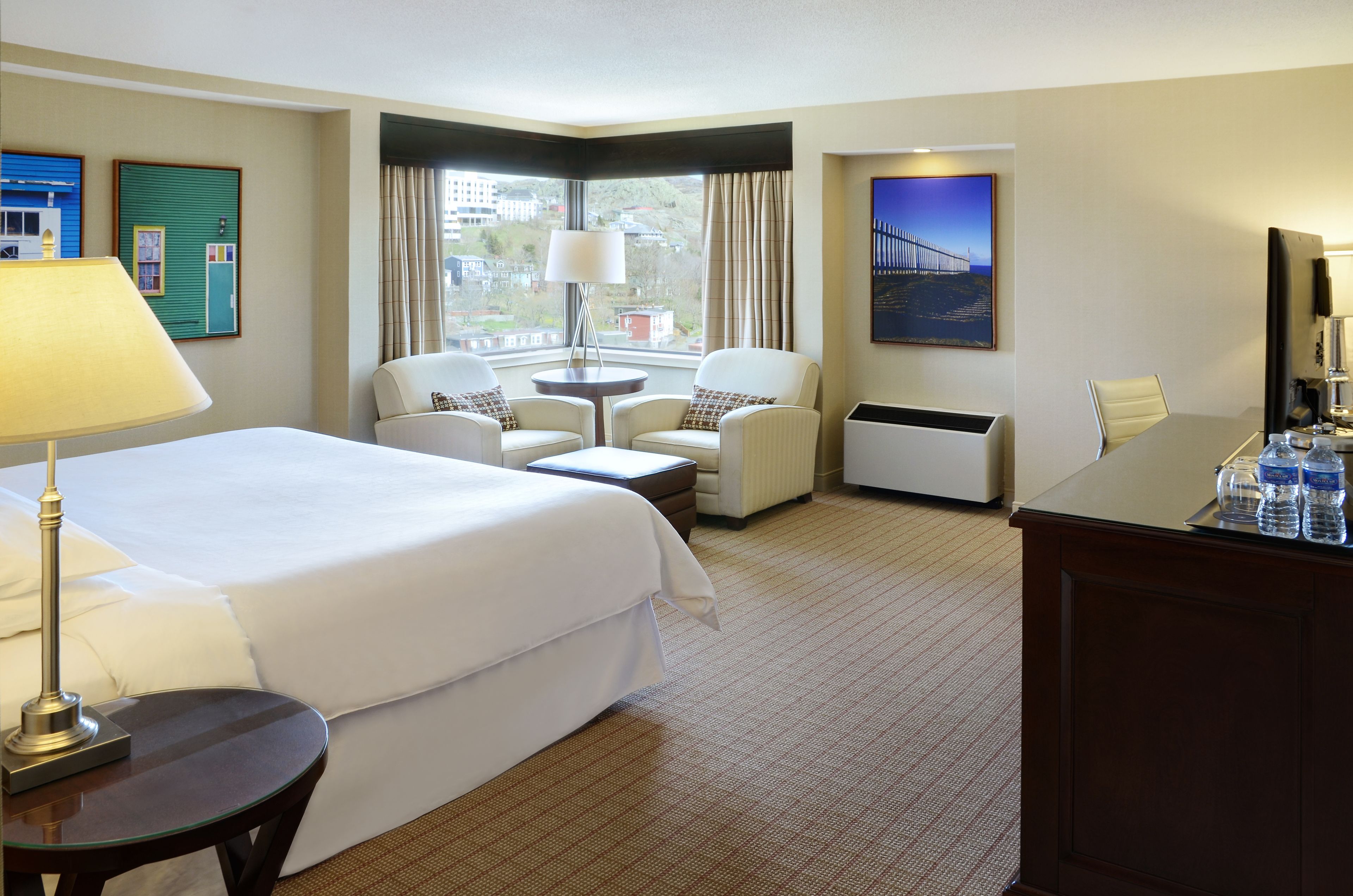 A hotel room with a large bed, two armchairs, a small table, a desk, and a view of a bridge through the window.
