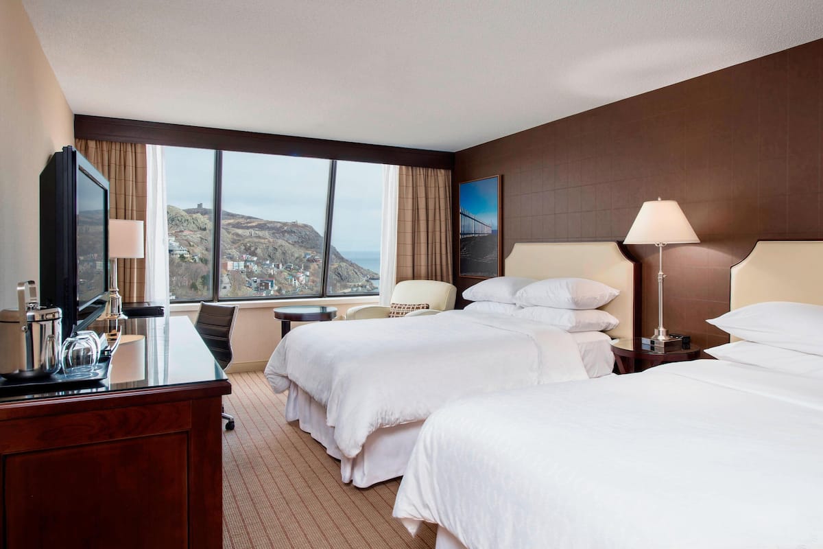 Ein Hotelzimmer mit zwei Betten, einem Schreibtisch mit Lampe, einem Sessel und Blick auf eine gebirgige Landschaft durch große Fenster.