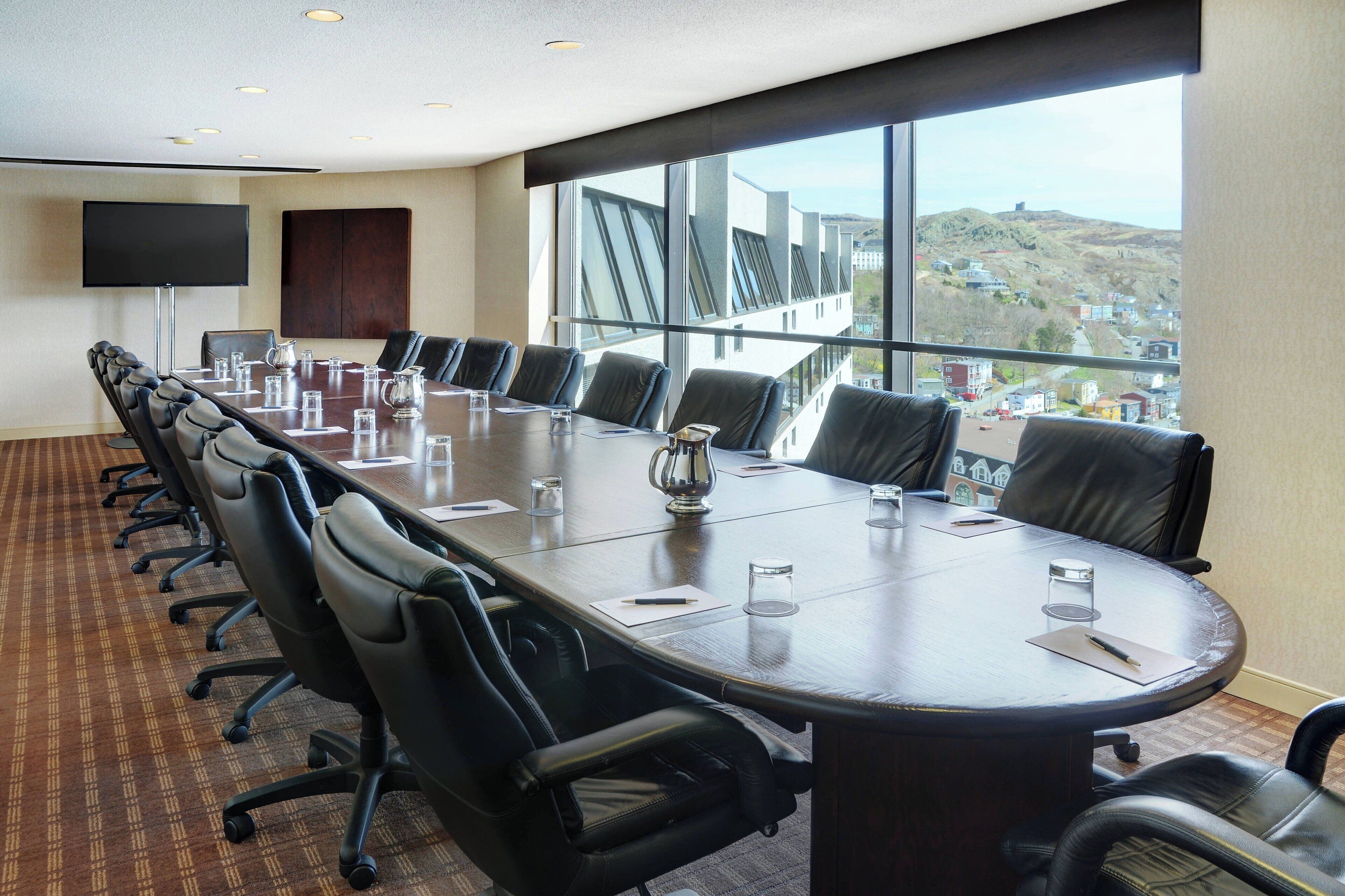 A conference room with a long table, multiple chairs, a TV, and large windows with a view of a town.