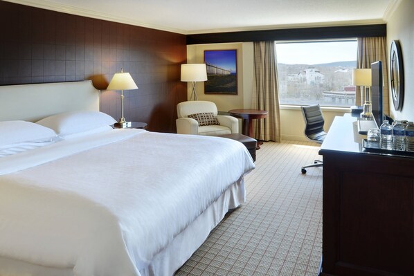 A hotel room with a large bed, two chairs, a desk, and a view of a bridge and buildings.