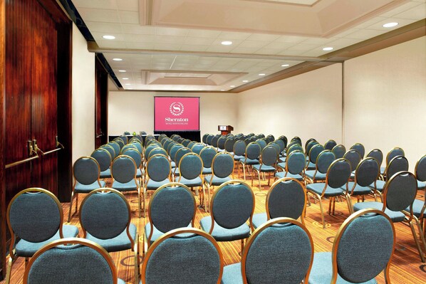 Une salle de conférence avec des rangées de chaises, un pupitre et un écran affichant le logo de la chaîne Sheraton.