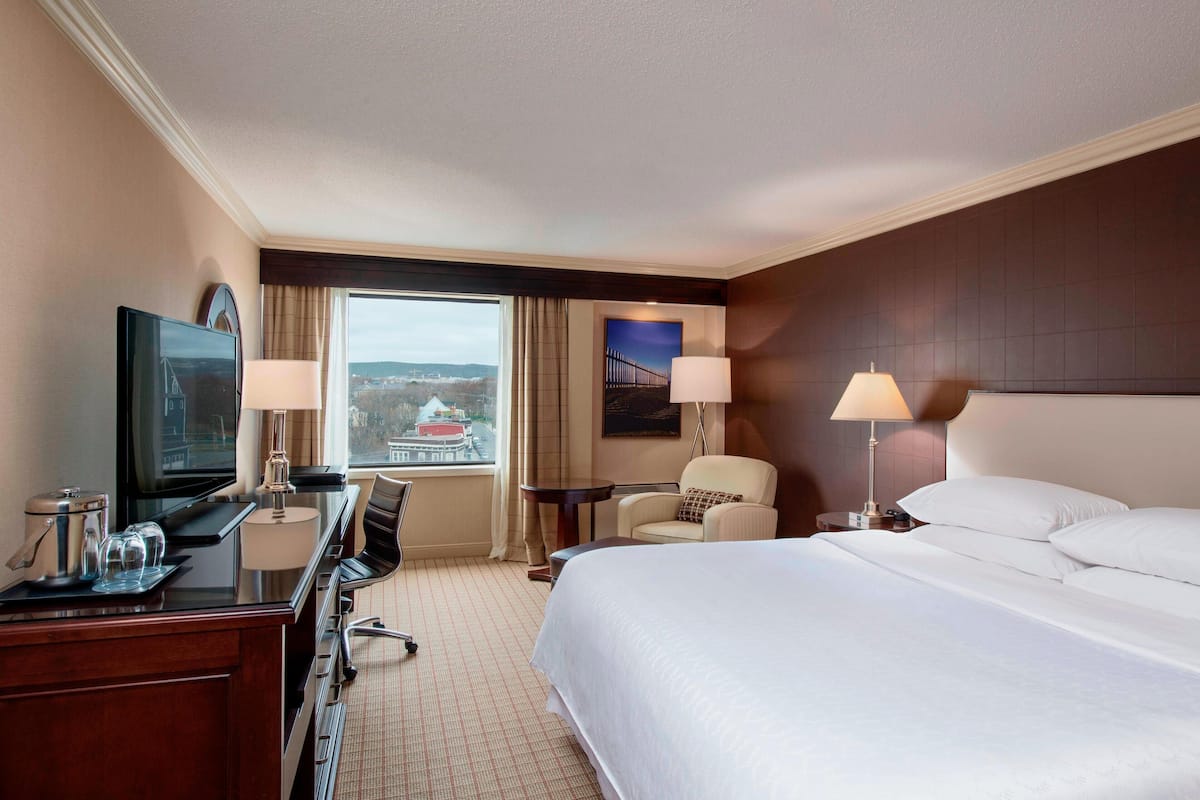 Ein Hotelzimmer mit einem Bett, einem Schreibtisch mit Computer, einem Sessel, einem Fernseher und einem Fenster mit Blick auf eine Brücke.