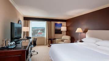 Ein Hotelzimmer mit einem Bett, einem Schreibtisch mit Computer, einem Sessel, einem Fernseher und einem Fenster mit Blick auf eine Brücke.