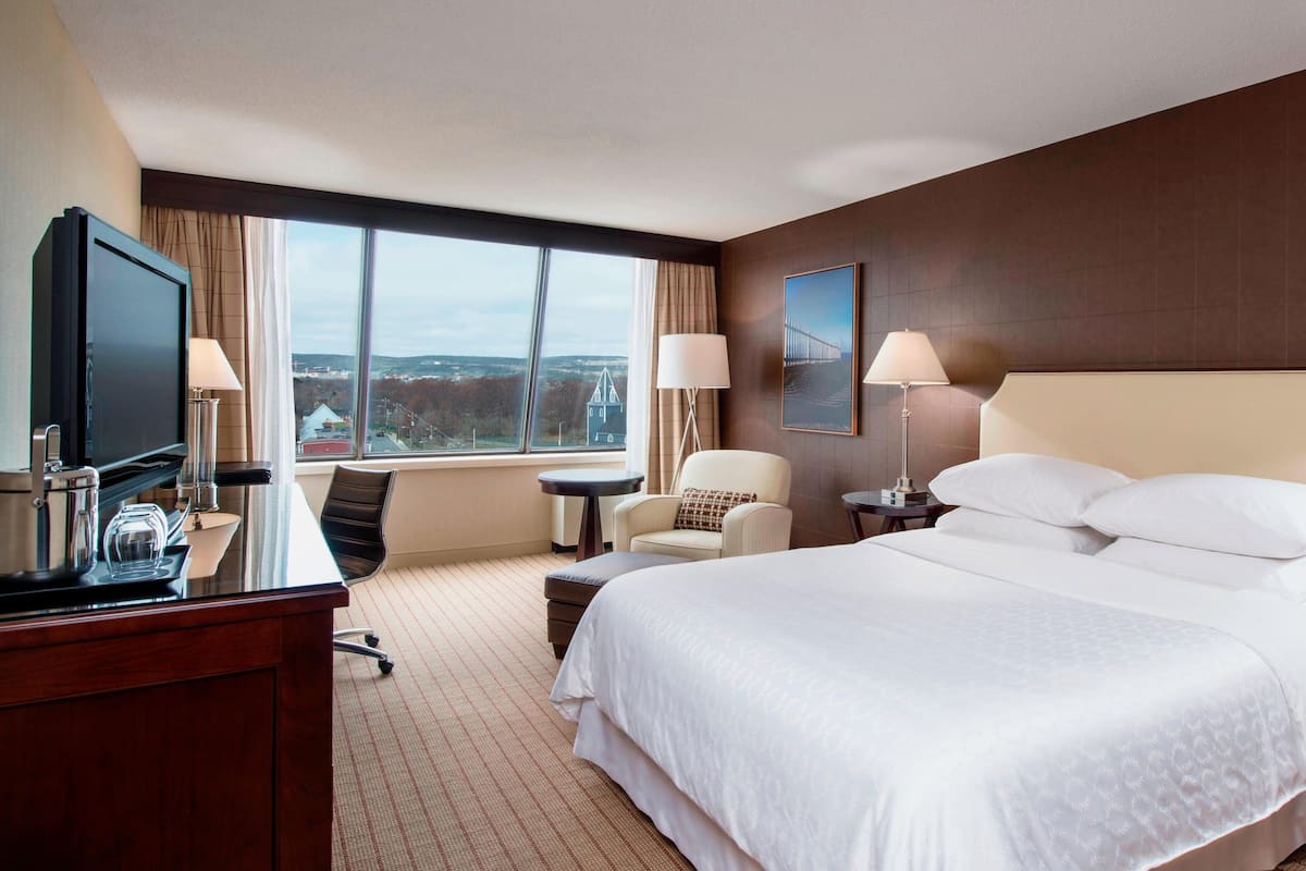 Ein Hotelzimmer mit einem großen Bett, einem Schreibtisch mit Fernseher, zwei Stühlen, einem kleinen Tisch und Blick auf eine Brücke sowie auf die Stadtlandschaft.