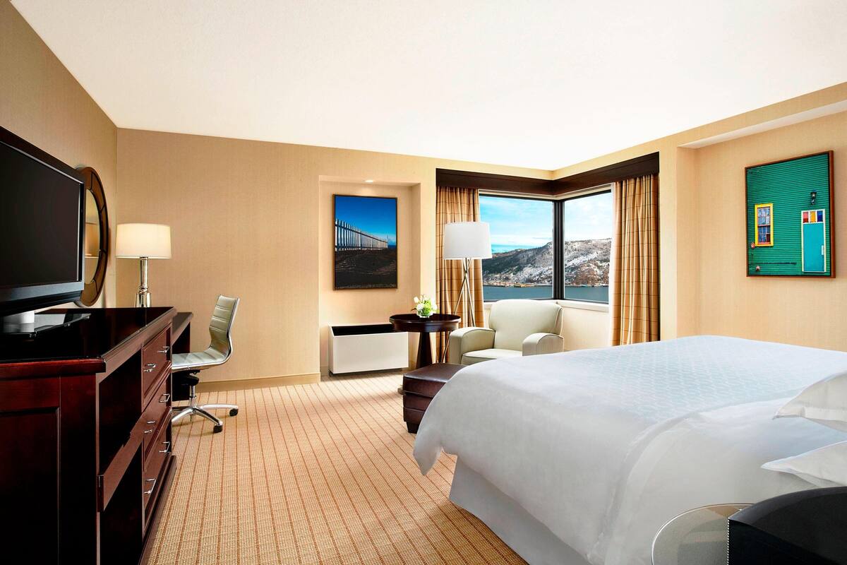 Ein Hotelzimmer mit einem großen Bett, einem Schreibtisch mit Stuhl, einem Fernseher und einem Fenster mit Blick auf eine Brücke und Berge.