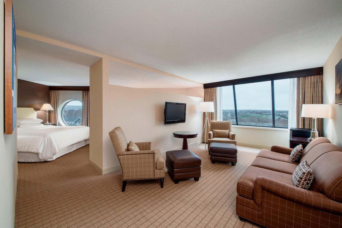 Ein Hotelzimmer mit einem Bett, zwei Sesseln, einem Sofa, einem Fernseher und einem großen Fenster mit Blick auf die Stadt.