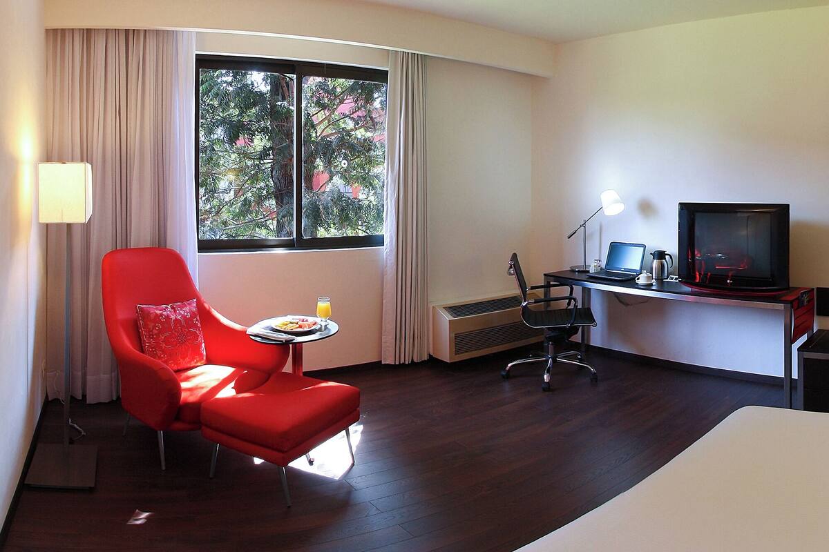 A hotel room with a red armchair, a small table with a plate of food and a glass of juice, a desk with a laptop, a television, and a heater.