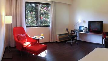 Habitación de hotel con un sillón rojo, una mesita con un plato de comida y un vaso de jugo, un escritorio con una computadora portátil, un televisor y un calefactor.