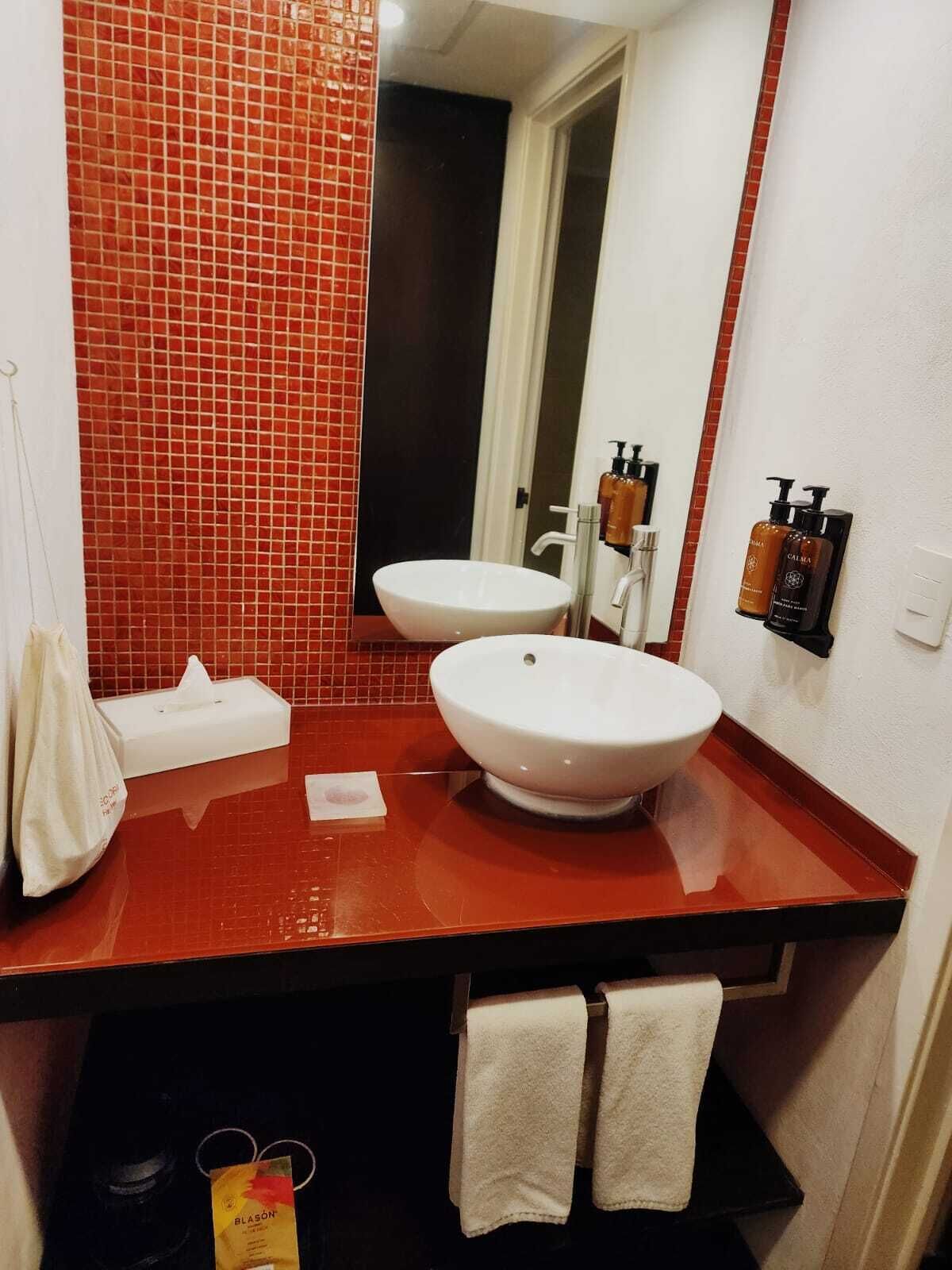 Un baño con pared de azulejos rojos, dos lavamanos, un espejo y toallas.