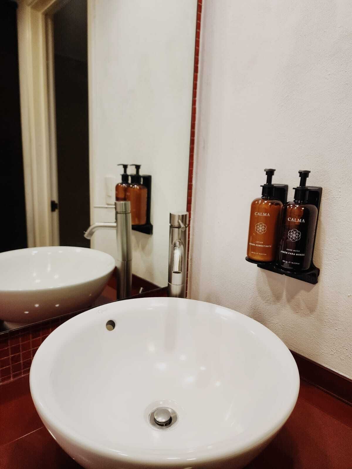 Un baño con un lavamanos blanco, un espejo y dos botellas de jabón de manos Calma sobre una repisa negra.