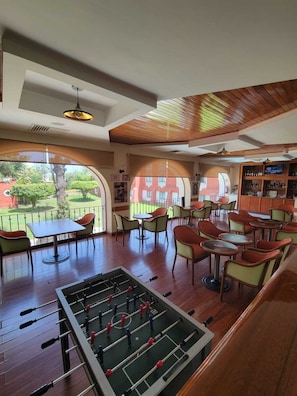 Una habitación con mesa de fútbol, varias mesas y sillas, y vista a un jardín.