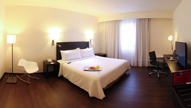 Habitación de hotel con una cama grande, mesitas de noche, un escritorio con silla, un televisor y una silla moderna.