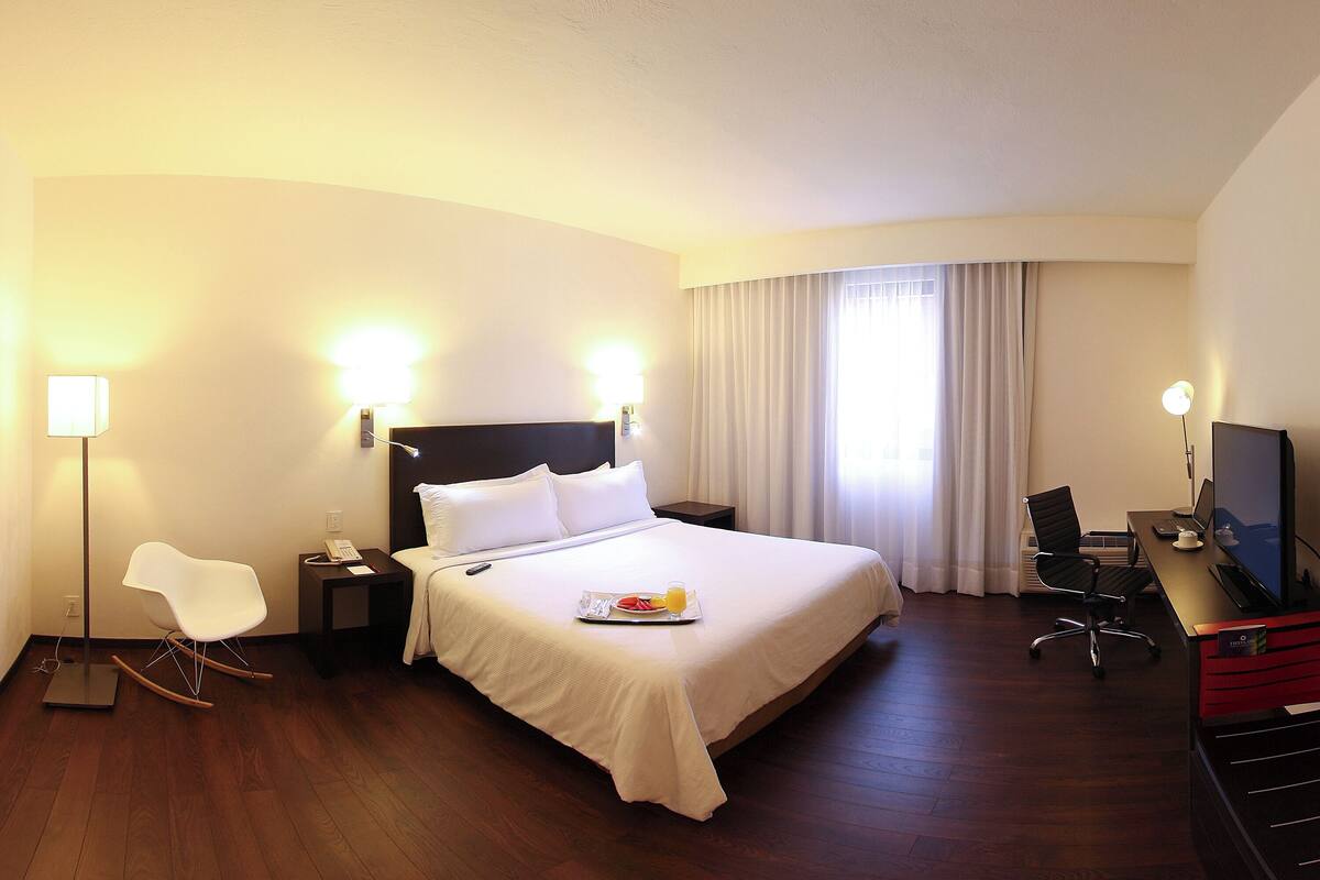 A hotel room with a large bed, bedside tables, a desk with a chair, a TV, and a modern chair.