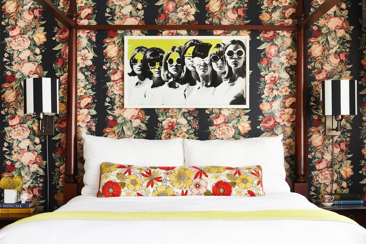 A bedroom with a floral wallpaper, a bed with white bedding, a colorful pillow, and a framed picture on the wall.