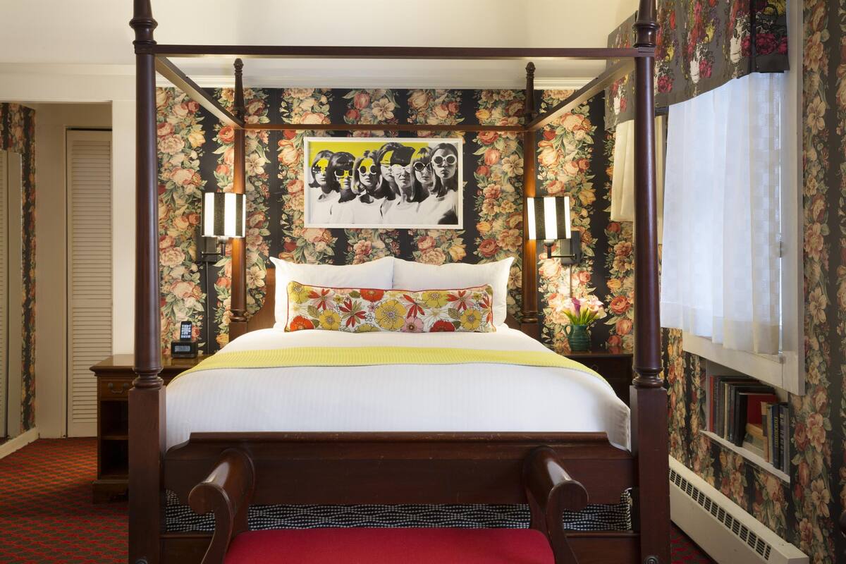 A four-poster bed with a floral-patterned headboard, a bedside table with a lamp, and a framed artwork above the bed.