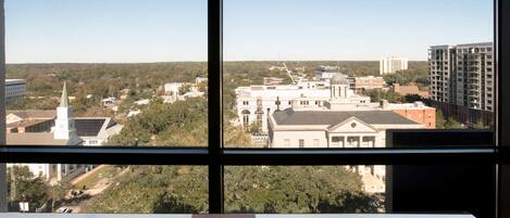 A room with a view of a cityscape through a window, featuring buildings and greenery.