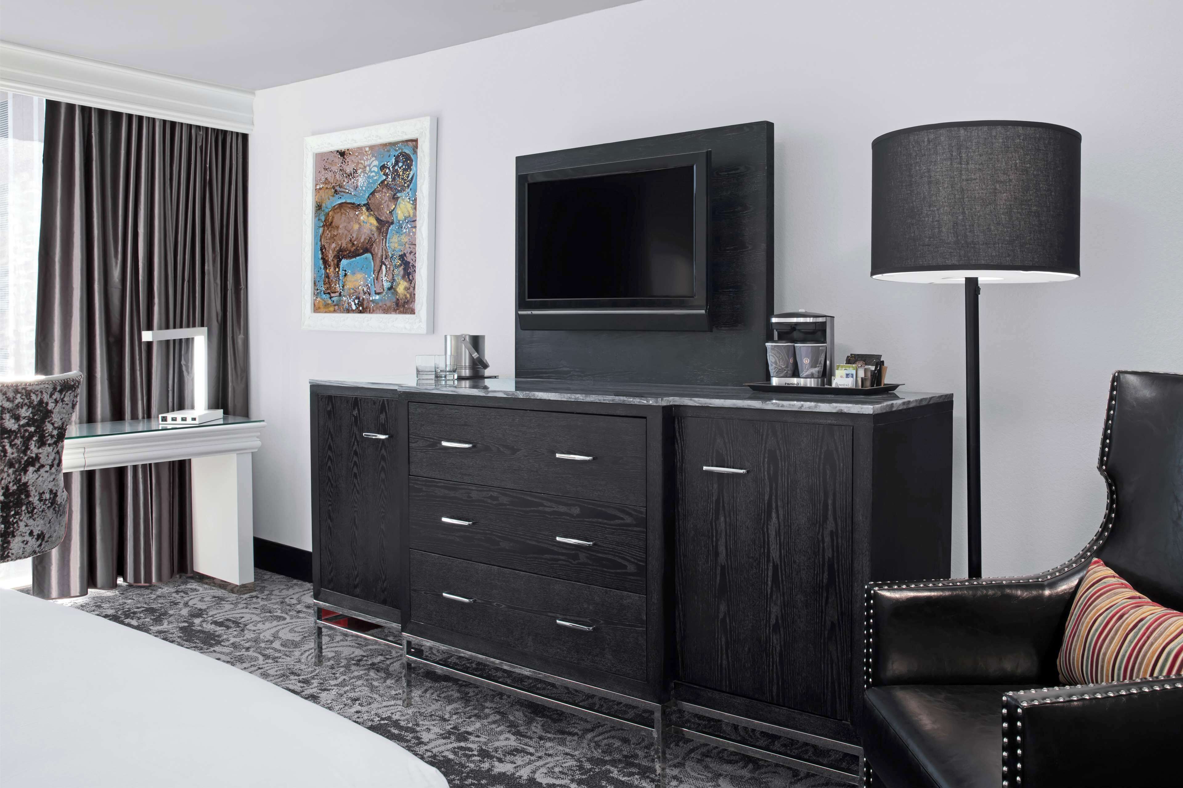 A modern hotel room with a large flat-screen TV mounted on a dark wooden cabinet, a black floor lamp, and a framed artwork on the wall.