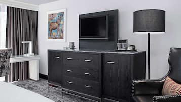 A modern hotel room with a large flat-screen TV mounted on a dark wooden cabinet, a black floor lamp, and a framed artwork on the wall.