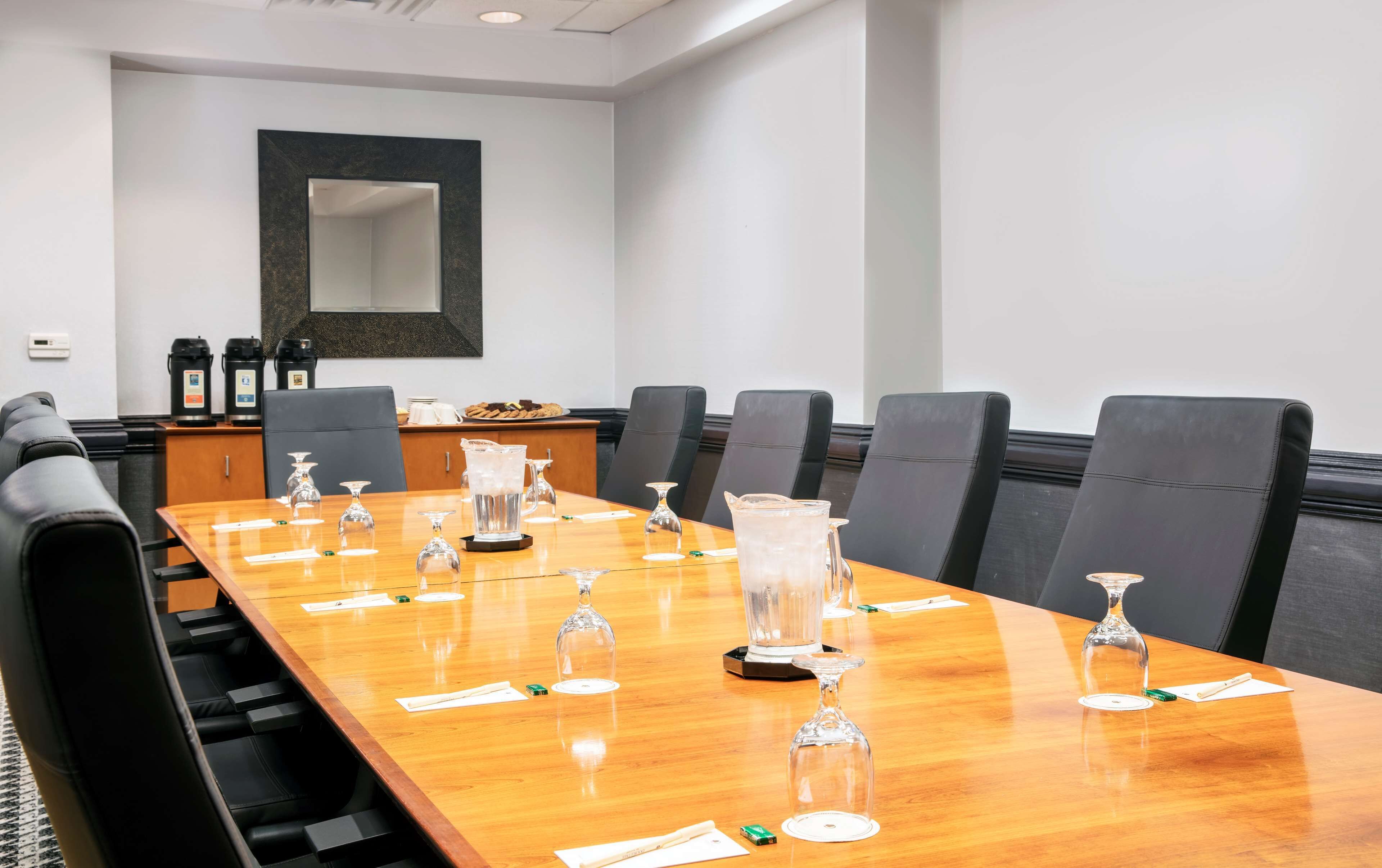 A conference room with a long wooden table, black leather chairs, and a large mirror on the wall.